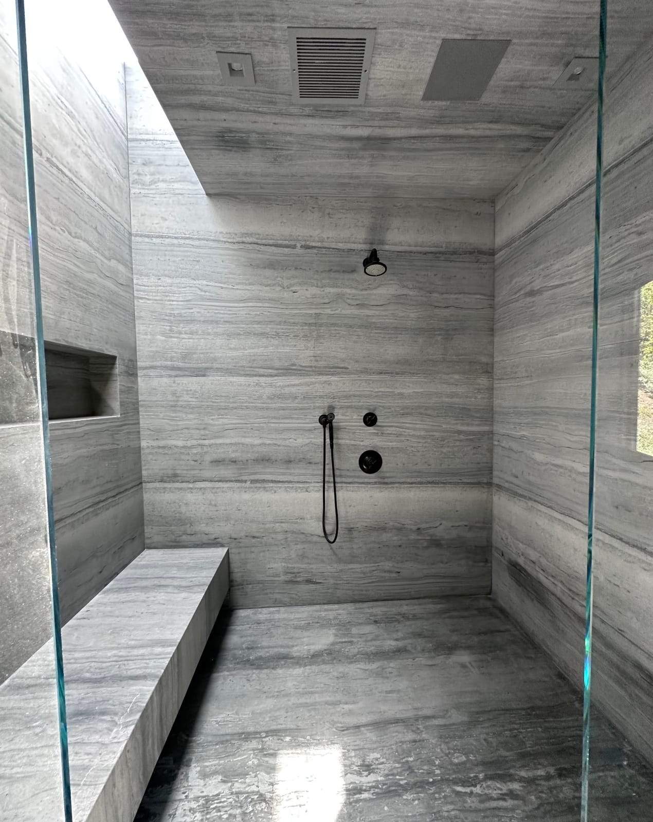 Remodeled walk-in shower clad in vein-cut stone, with a floating bench and frameless glass
