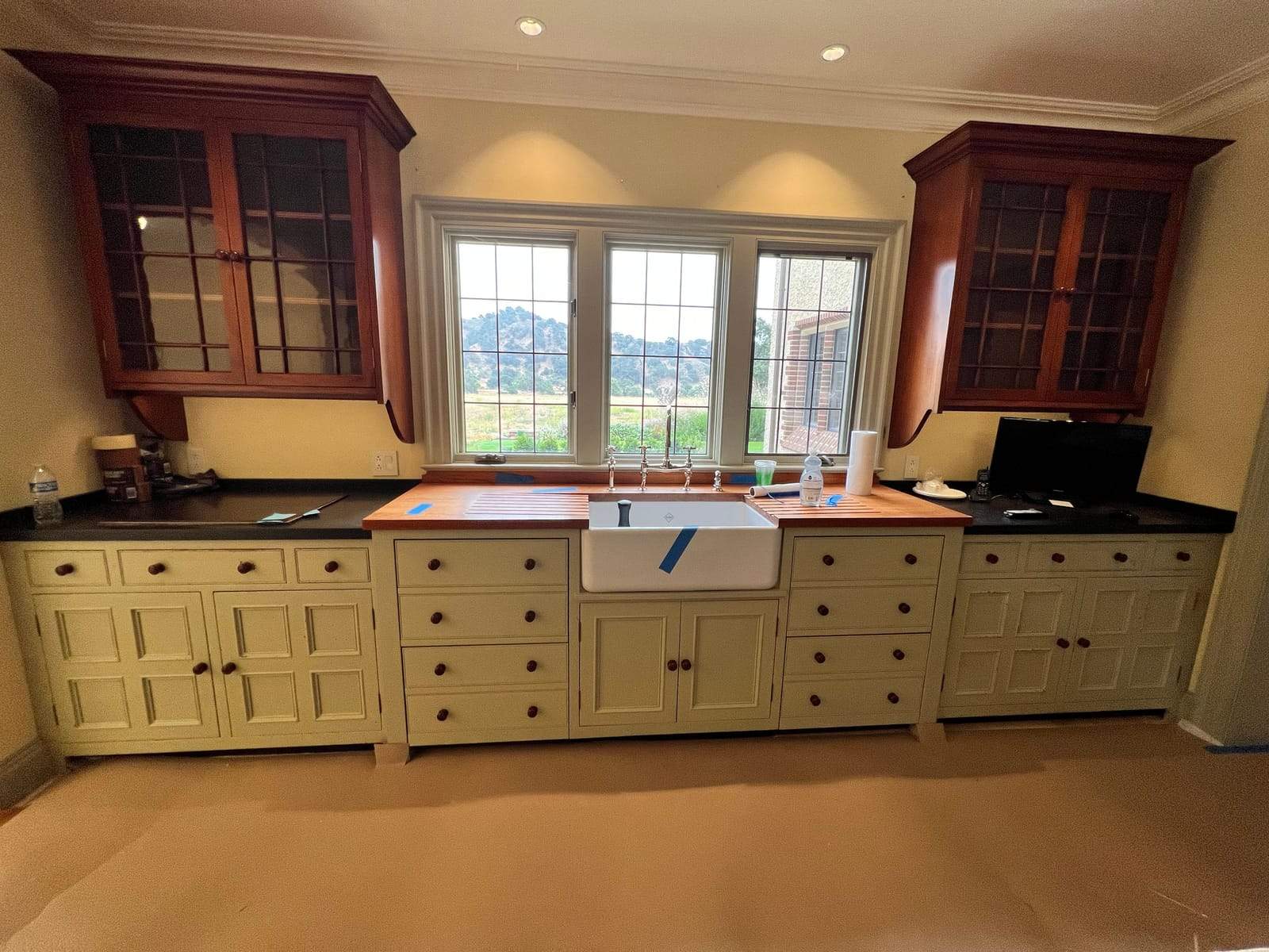 Kitchen sink wall with cream cabinetry, cherry glass-front uppers, and a farmhouse sink under leaded windows
